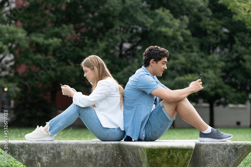 Teenager couple sitting back to back and using phone outdoors in a park. Digital and social media addiction concept.