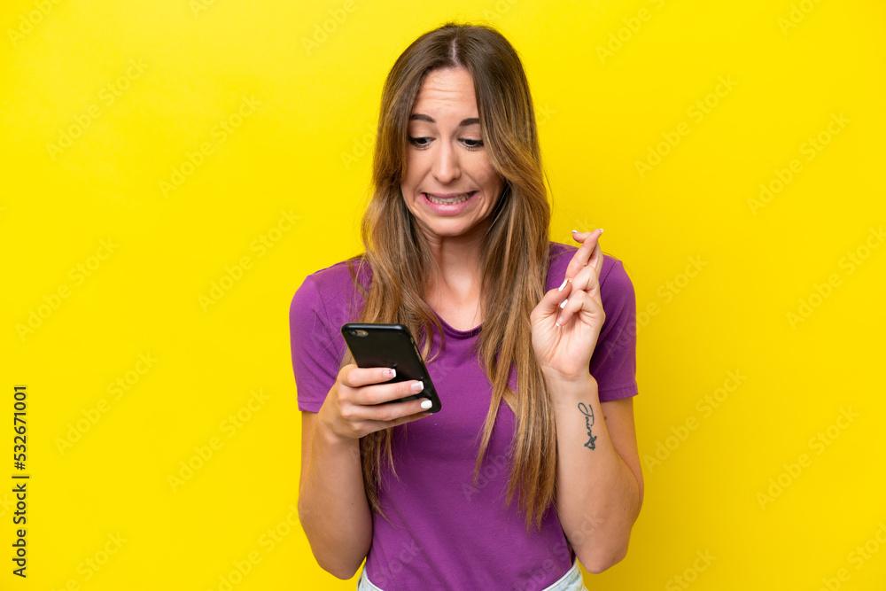 Young caucasian woman isolated on yellow background using mobile phone with fingers crossing Young caucasian woman isolated on yellow background using mobile phone with fingers crossing