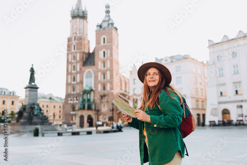 Attractive young female tourist is exploring new city. Redhead girl holding a paper map on Market Square in Krakow. Traveling Europe in autumn. St. Marys Basilica © mdyn