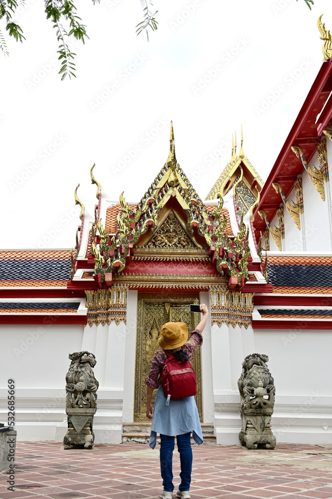 A women tourist wearing a yellow hat and jeans uses a mobile phone to take a selfie as she walks through the beauty of pagodas and ancient architecture inside Wat Pho or Recling Buddha Temple in Bangk