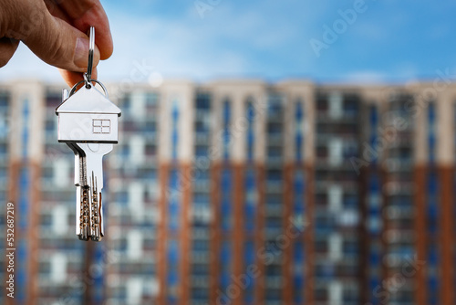 The keys to the lock with a keychain in the form of a house in the hand of a man against the background of a multi-storey residential building. Purchase, rent, insurance of a house, apartment.