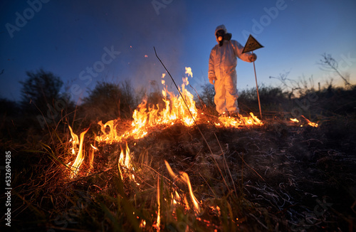 Fireman ecologist working in field with wildfire at night. Man in protective radiation suit and gas mask near burning grass with smoke, holding warning sign. Natural disaster concept.
