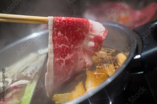 Hand using chopsticks pick premium raw beef sliced beef over the hot pot soup for Shabu Shabu or Sukiyaki dipping and dropping. Popular food is cooked piece by piece by the diner, served with dipping 