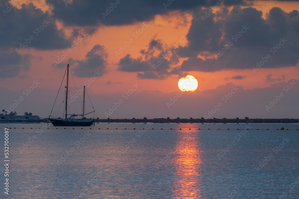 Sunrise over the Mediterranean sea, on a cloudy summers morning.  The sun is reflecting on the calm sea, on which there are a few sailing boats