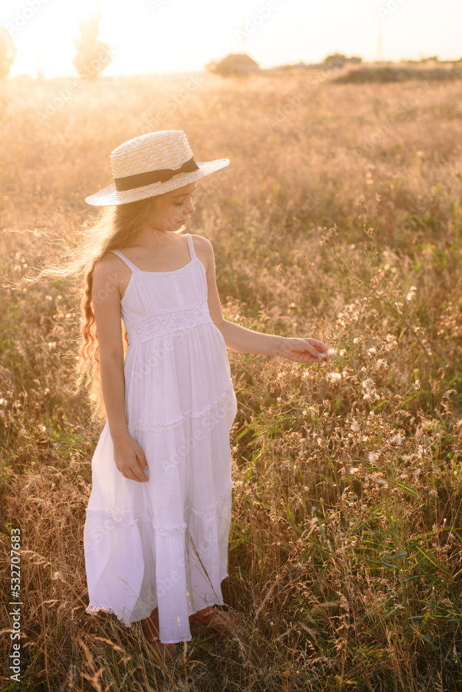 Cute little girl with blond long hair in a summer field at sunset with a white dress with a straw hat