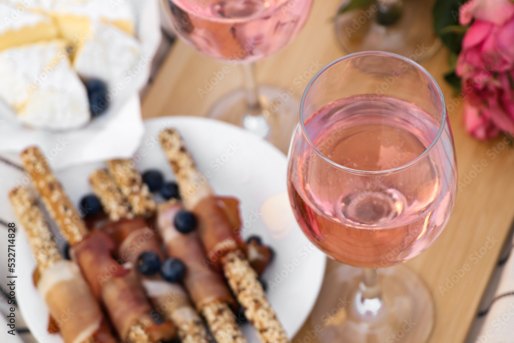 Glasses of delicious rose wine, flowers and food on wooden board, closeup