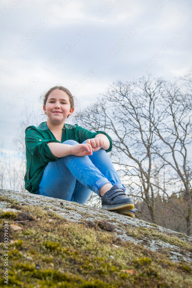 In the spring afternoon, a girl with a scythe sits on the edge of a cliff in blue jeans and a green shirt, side view.