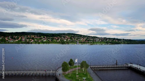 Aerial view over a lake. Flying backwards revealing the pier with a landing stage. People walk across the mooring place on a summer evening overlooking a town with red cottages against the mountain.  photo