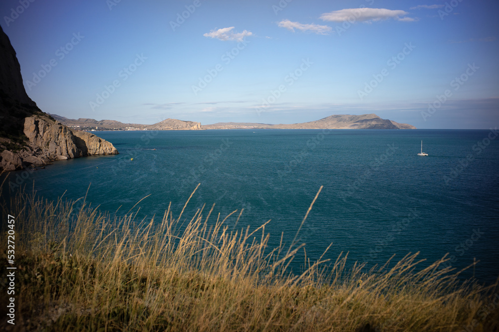 view from the shore to the endless sea, dry autumn grass and a small boat in the distance. high mountain on the left.