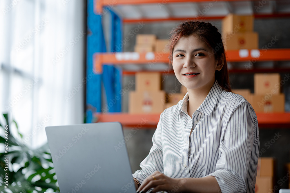 Fototapeta premium A beautiful Asian business owner opens an online store, she is checking orders from customers, sending goods through a courier company, concept of a woman opening an online business.