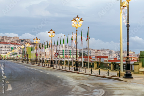 Algiers city empty Che Guevara Boulevard Street Road with white buildings, Algerian flags and light poles, road panel signs, cars from far and cloudy sky. 