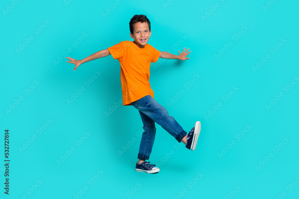 Full length photo of cheerful boy dressed trendy orange clothes stand one leg foot good mood empty space isolated on cyan color background Full length photo of cheerful boy dressed trendy orange clothes stand one leg foot good mood empty space isolated on cyan color background