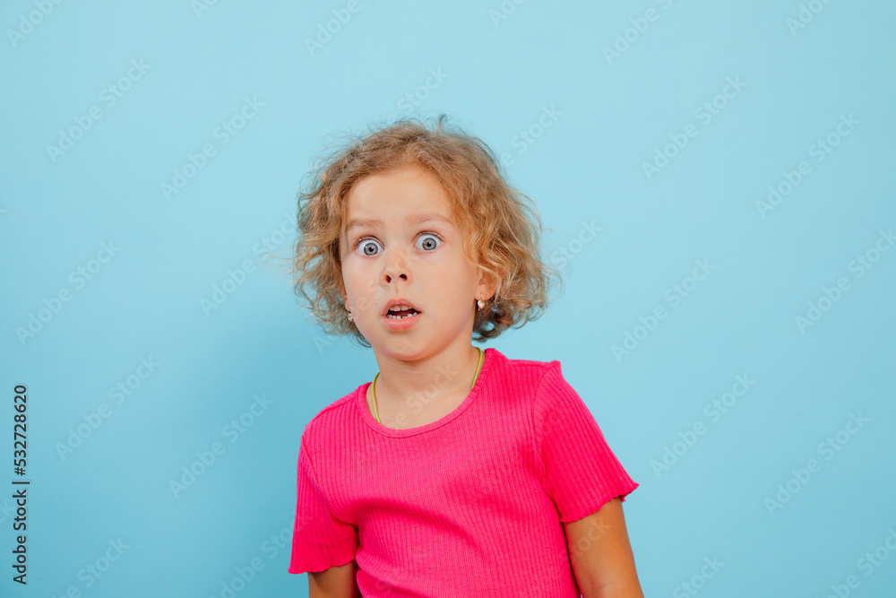 Portrait of amazed little girl with curly fair hair wearing pink T-shirt, looking with open mouth on blue background. Portrait of amazed little girl with curly fair hair wearing pink T-shirt, looking with open mouth on blue background.
