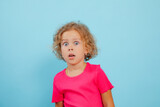 Portrait of amazed little girl with curly fair hair wearing pink T-shirt, looking with open mouth on blue background.