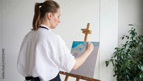 Mature woman artist with ponytail paints sky landscape on canvas. Skilled lady master in white enjoys painting picture on wooden easel closeup