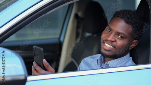 African American driver sitting at car steering wheel talks with friends via videocall. Bearded man spends time in traffic jams communicating with friends