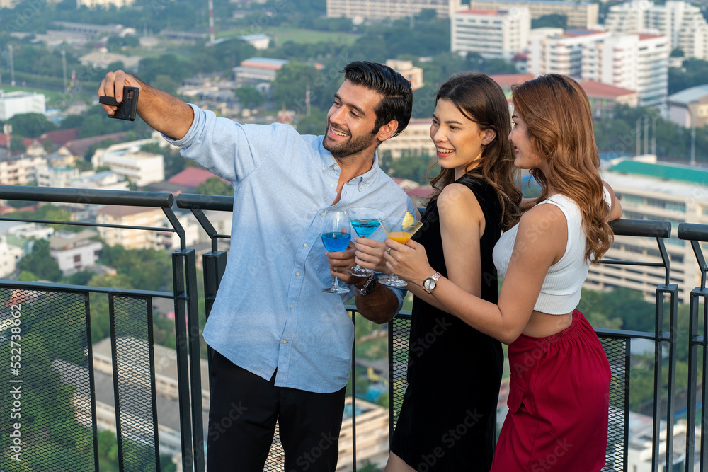 Obraz premium International group of male and female friends holding a smartphone selfie with a girl friend holding a glass of cocktails, party and restaurant on the roof of the building.