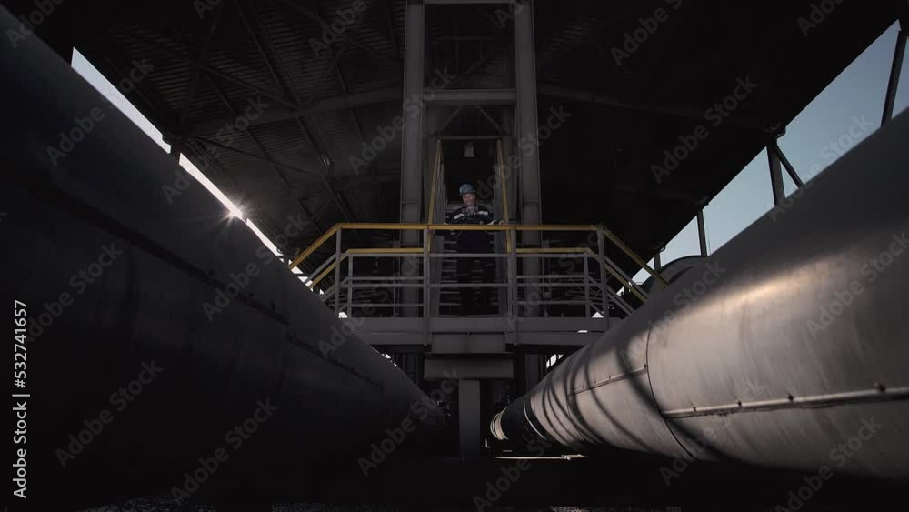 Oil and gas worker walks down an iron ladder in front of a large oil pipeline. Oil and gas worker walks down an iron ladder in front of a large oil pipeline.