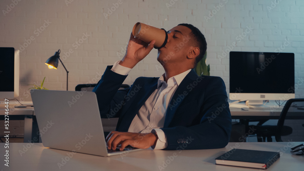 African american manager in suit drinking coffee to go and using laptop in office at night. African american manager in suit drinking coffee to go and using laptop in office at night.