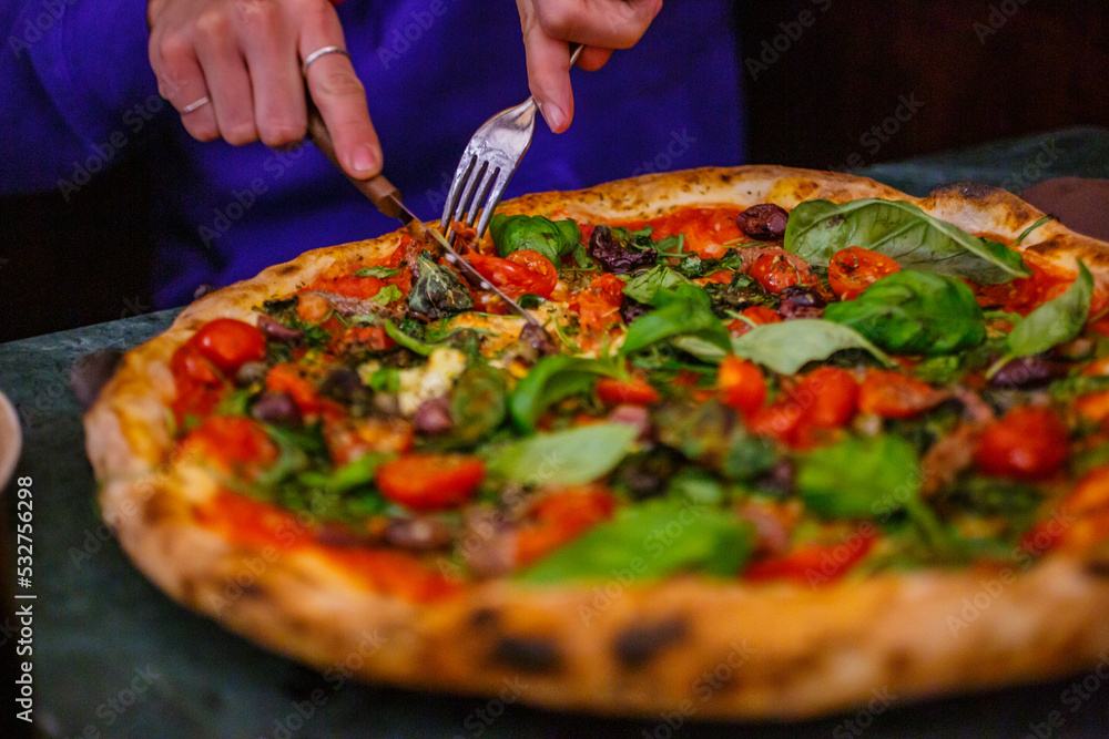 Pizza napoletana con pomodorini, basilico fresco, aglio, origano e olio mentre viene tagliata con forchetta e coltello da un cliente