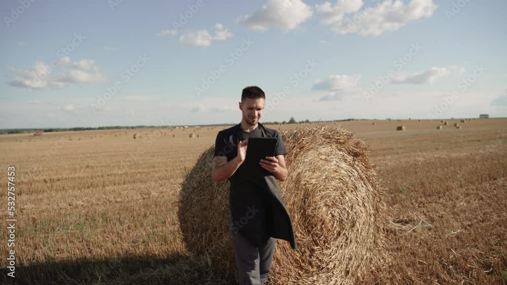 Farmer stands with tablet on agricultural field near sheaf of hay. Farmer stands with tablet on agricultural field near sheaf of hay.