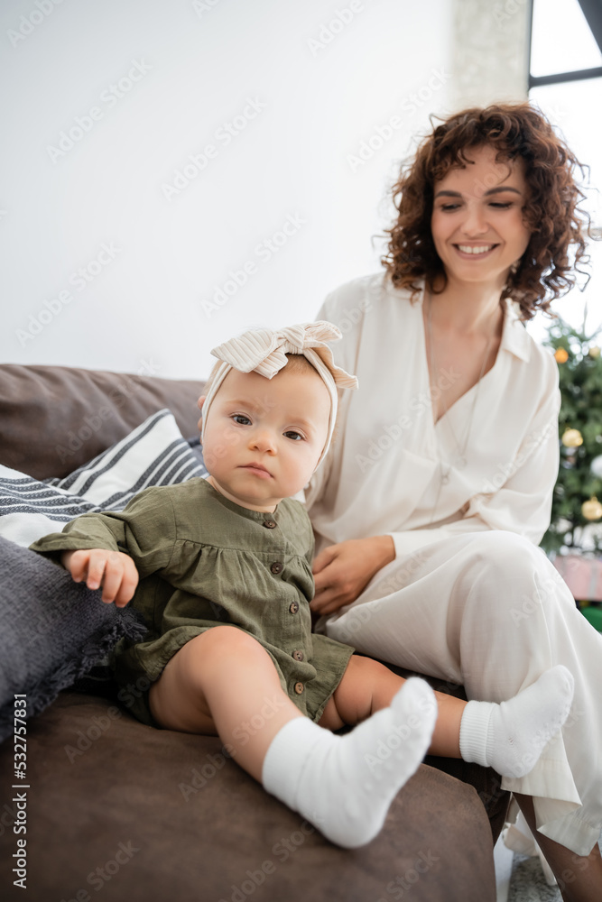 happy mother in dress smiling while looking at infant daughter sitting on sofa. happy mother in dress smiling while looking at infant daughter sitting on sofa.