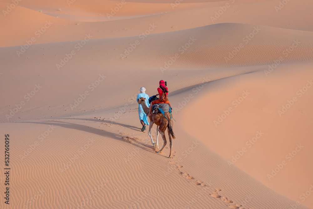 A woman in a red turban riding a camel across the thin sand dunes of the in Western Sahara Desert, Morocco, Africa