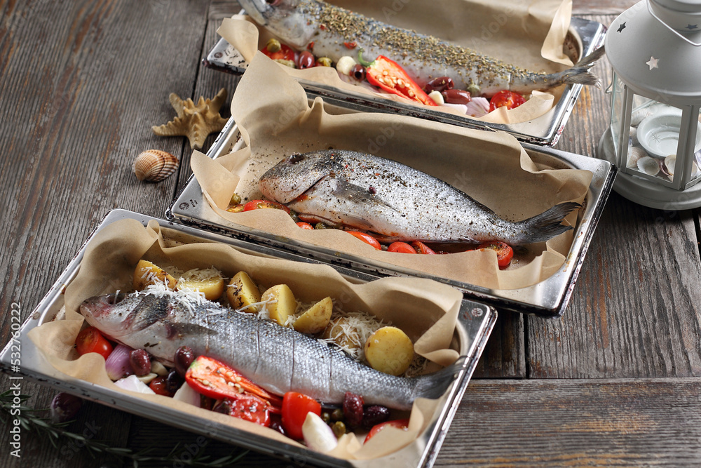 Sea bass, sea bream and mackerel in baking dishes, on table. Baking fish with vegetables in sheet pans.