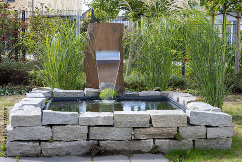 Urban garden with a square high pond and a small waterfall created by a stainless steel outlet which is mounted on a wooden stele, surrounded by green grass