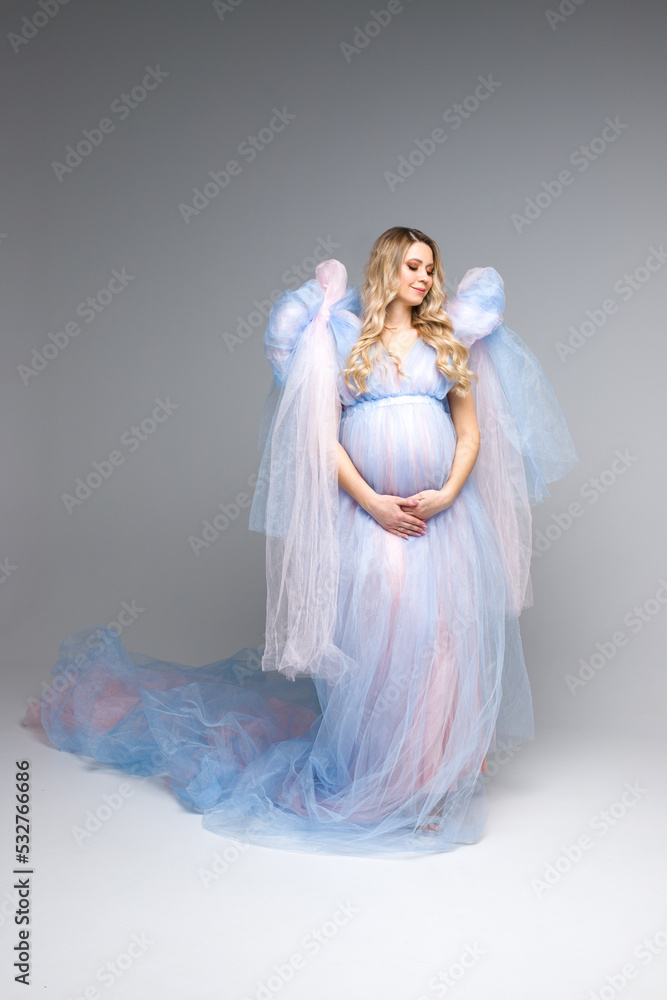 A happy pregnant girl with blonde hair in a blue airy dress on a gray background. Beautiful pregnant girl in a blue dress. Studio shooting. Vertical photo.