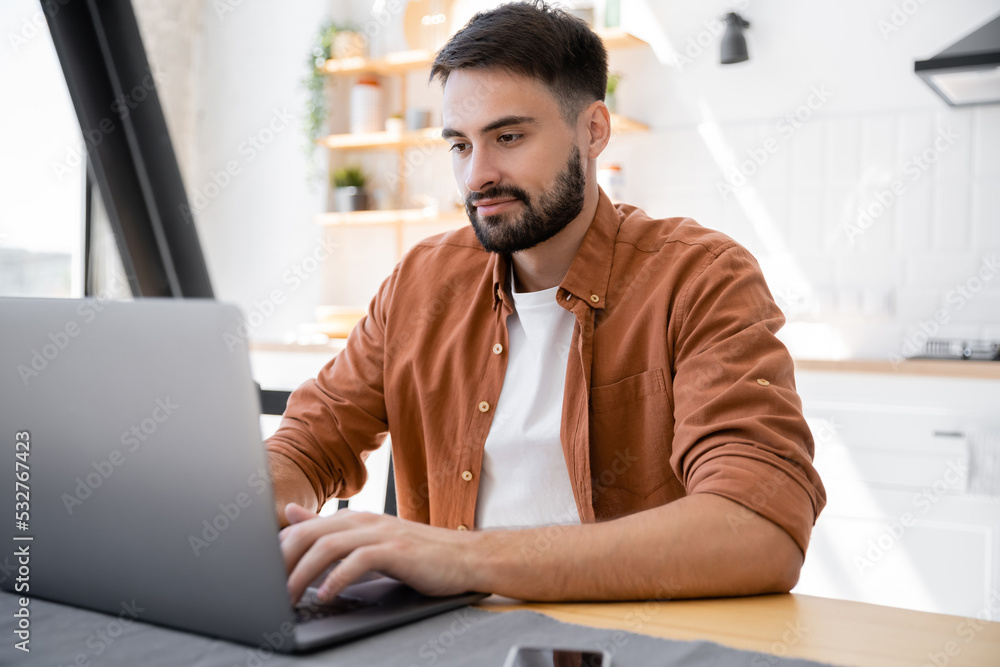Obraz premium bearded freelancer using laptop while working from home near smartphone on table.