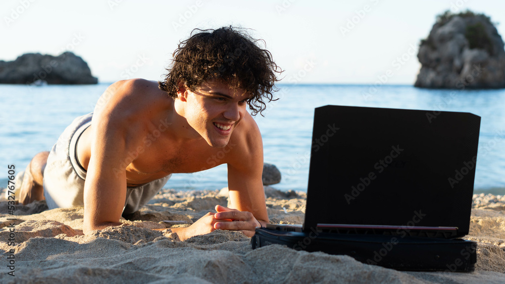 Young attractive man doing plank at the beach follow his trainer via laptop with the sea at the background. Workout exercise. Healthy handsome active man with muscular body doing plank. 