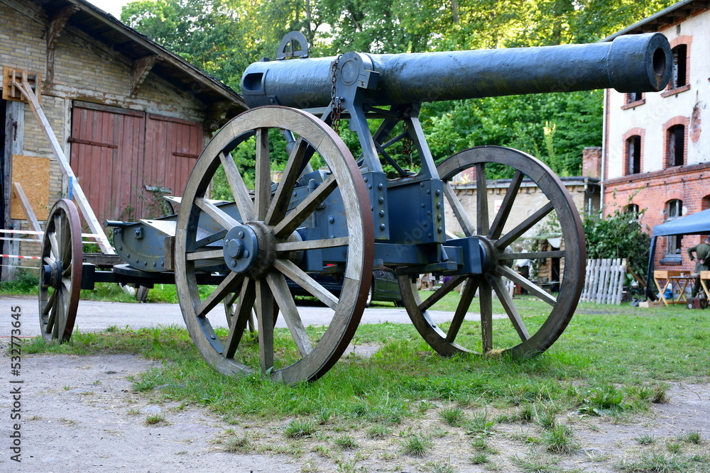 Single massive grey cannon replica on two rusty wheels with base and barrel visible stanting next to an ad hoc wooden barricade in the middle of a lush field, pastureland, or meadow seen in Poland