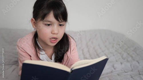 Close up of little girl in pink sweatshirt laying on the bed reading book aloud with open mouth. Child is turning the pages with an interest. Study at home, Homeschooling concept and cozy leisure time