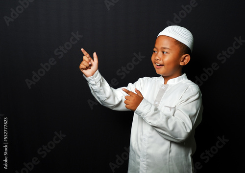 An Asian kid wearing a cap points his hand to the empty space isolated on black background.