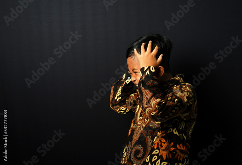Asian kid wearing Batik holding his head and looks frustrated isolated on black background. photo