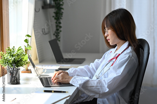 Asian female doctor working with laptop while wearing white medical uniform at workplace.