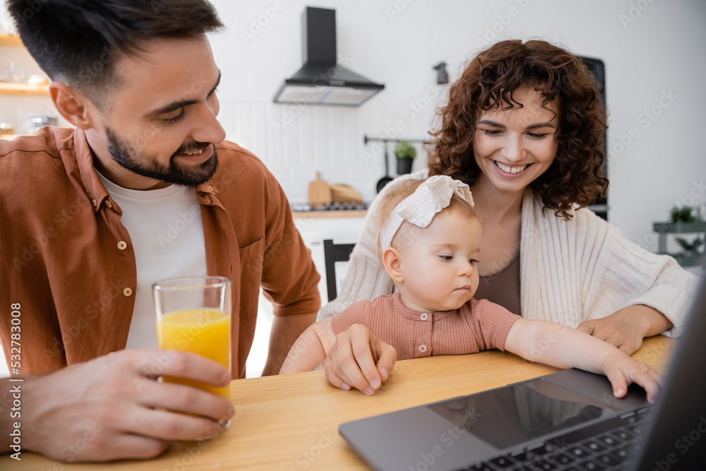 cheerful man holding glass of orange juice near wife and infant daughter using laptop. cheerful man holding glass of orange juice near wife and infant daughter using laptop.