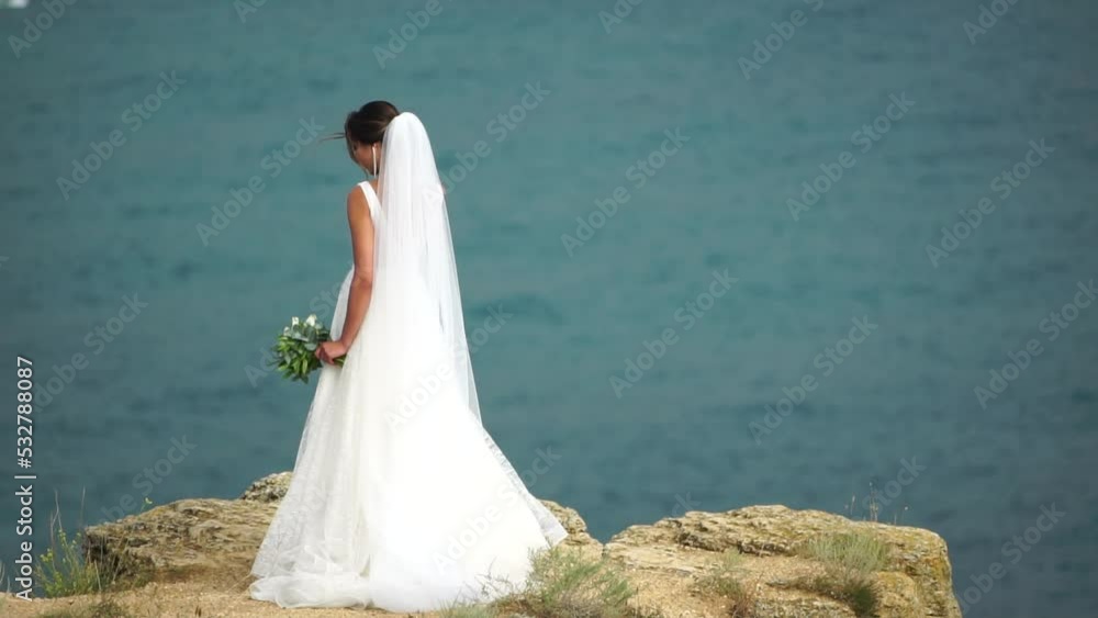 A beautiful bride stands with her back against the background of the sea with a bouquet of flowers. A woman in a long white dress. A beautiful bride stands with her back against the background of the sea with a bouquet of flowers. A woman in a long white dress.