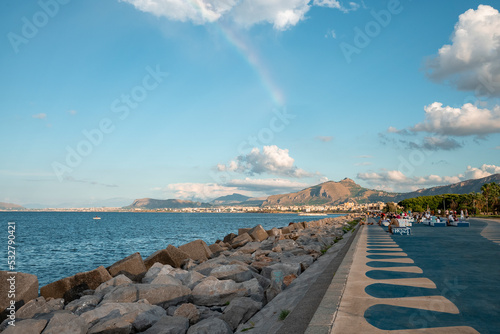 Distant view of tourists relaxing on promenade. Mountains and old town at coastline in background. Rocks arranged at seafront with blue sky in background on sunny day. photo