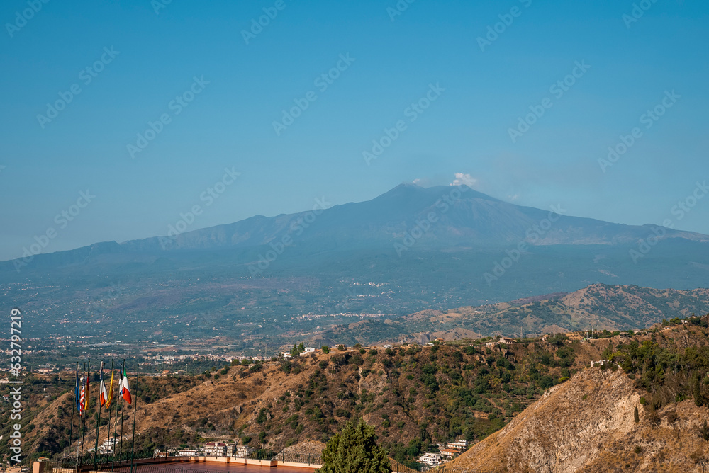 Beautiful Mount Etna amidst dramatic landscape with blue sky in background. Luxurious hotel Elios with flags in foreground. Scenic view of famous tourist attraction during sunny day.