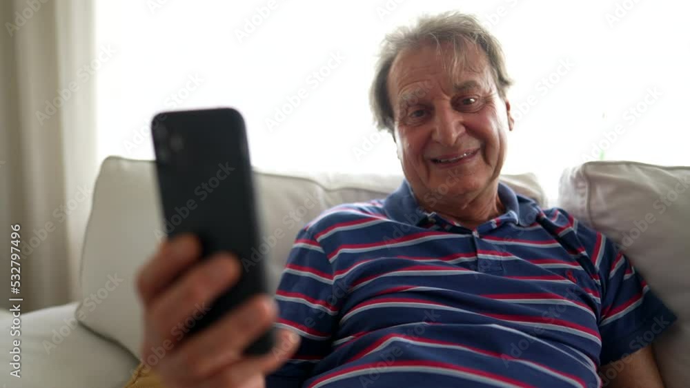 Senior man holding phone speaks in video with smartphone. Casual authentic older mature person using modern technology device in living room couch Senior man holding phone speaks in video with smartphone. Casual authentic older mature person using modern technology device in living room couch