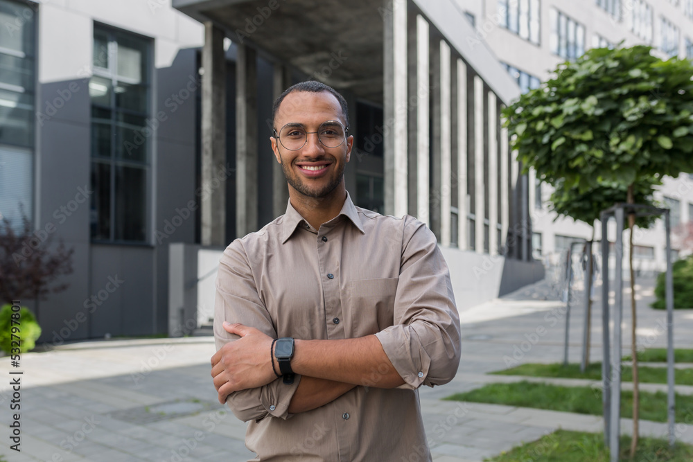 Young successful businessman smiling and looking at camera with crossed arms, man outside office building wearing shirt and glasses, startup investor. - obrazy, fototapety, plakaty 