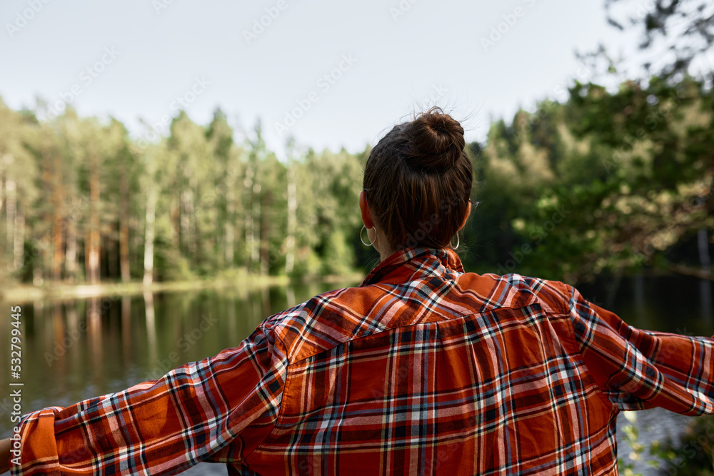 Rare view of woman in red checkered shirt outstretching hands admiring beauty of calm lake and coniferous forest on opposite side of river, enjoying tranquil and peaceful wild nature Rare view of woman in red checkered shirt outstretching hands admiring beauty of calm lake and coniferous forest on opposite side of river, enjoying tranquil and peaceful wild nature