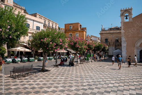 Taormina, Sicily, Italy. August 26, 2022. Male and female tourists exploring old townsquare on sunny day. Ancient belltower and historic buildings at coastal city with blue sky in background. photo