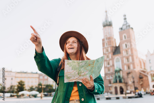 Attractive female tourist is exploring new city. Redhead woman pointing finger and holding a paper map on Market Square in Krakow. Traveling Europe in autumn. St. Marys Basilica. Active lifestyle
