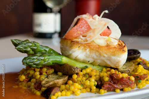 Seared halibut fillet on asparagus served with saffron-infused couscous, fingerling potatoes and sun dried tomatoes in sauce, and topped with grapefruit and thinly sliced bulb fennel. Horizontal. 