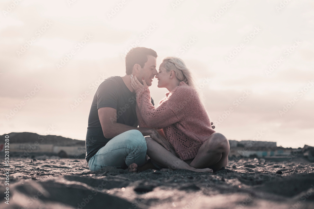 Romantic evening with girl and boy together sitting at the beach on the sand kissing in tenderness. Two people male and female in relationship sitting on the sand and enjoying sunset. Man woman kiss