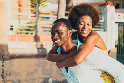 Happy black people couple with young man and woman having fun together in piggyback carrying activity. Two person african male and female enjoying leisure. Buildings houses in background. Love friends