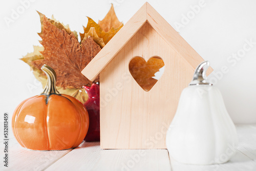 Autumn house insurance, sale, rent concept. White decorative pumpkins and autumn yellow leaves with miniature wooden house close-up and copy space..